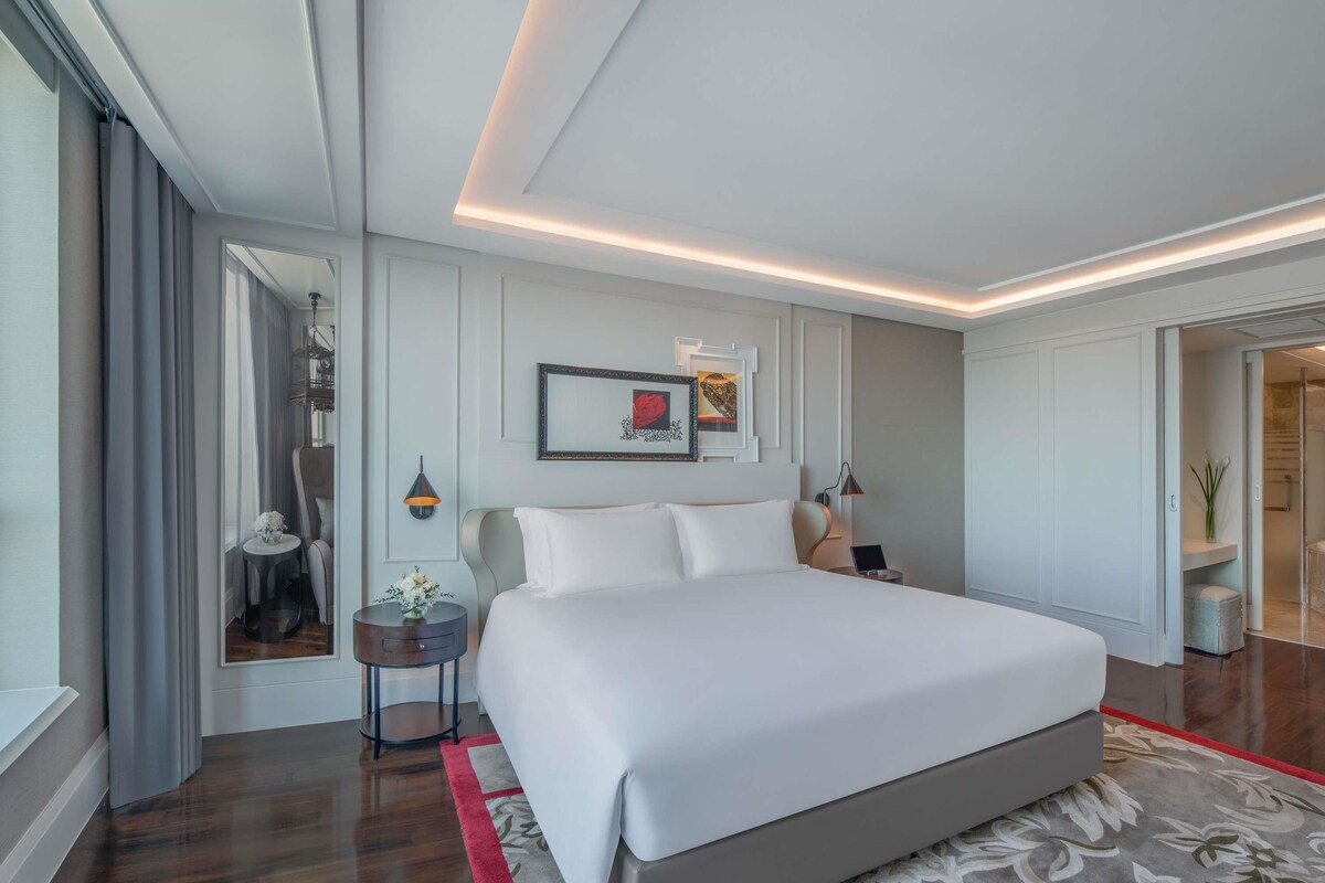 A modern hotel room with a large bed, a round side table, a mirror, and a door leading to a bathroom.