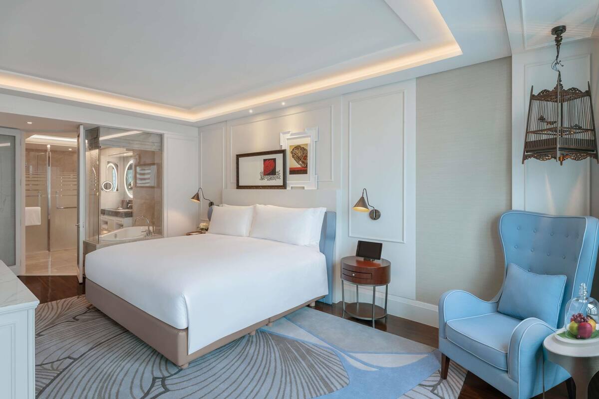 Une chambre d’hôtel moderne avec un grand lit, un coin salon comprenant un fauteuil bleu et une petite table d’appoint, ainsi qu’un espace douche doté de portes en verre.