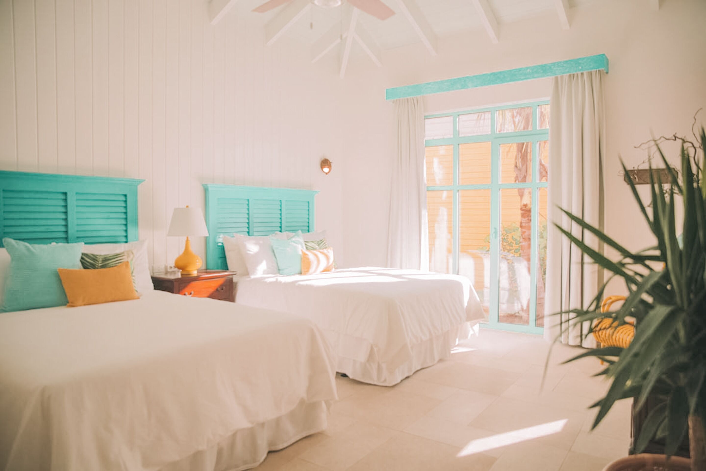 A bright, airy bedroom with two beds, teal accents, and a large window.