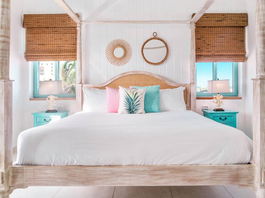 Una cama con dosel de madera, ropa de cama blanca y almohadas decorativas. Hay dos mesitas de noche con cajones azul verdosos y lámparas. La habitación tiene una ventana con vista a palmeras y edificios.