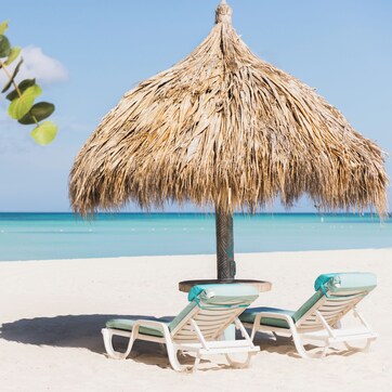 A beach scene with two straw umbrellas and two lounge chairs.