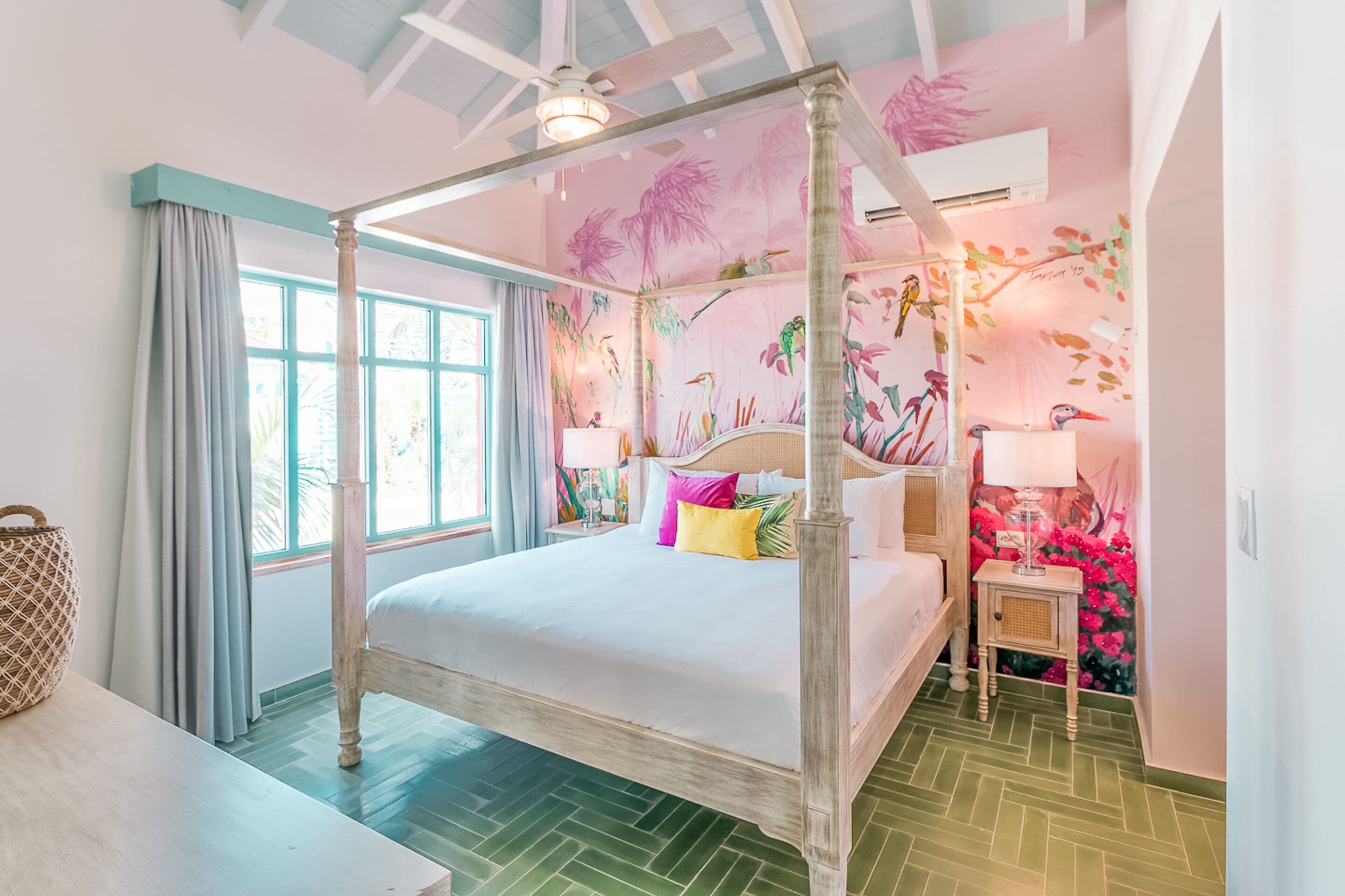 A four-poster bed with a white canopy, a small bedside table with a lamp, and a wall mural of tropical birds and flowers.