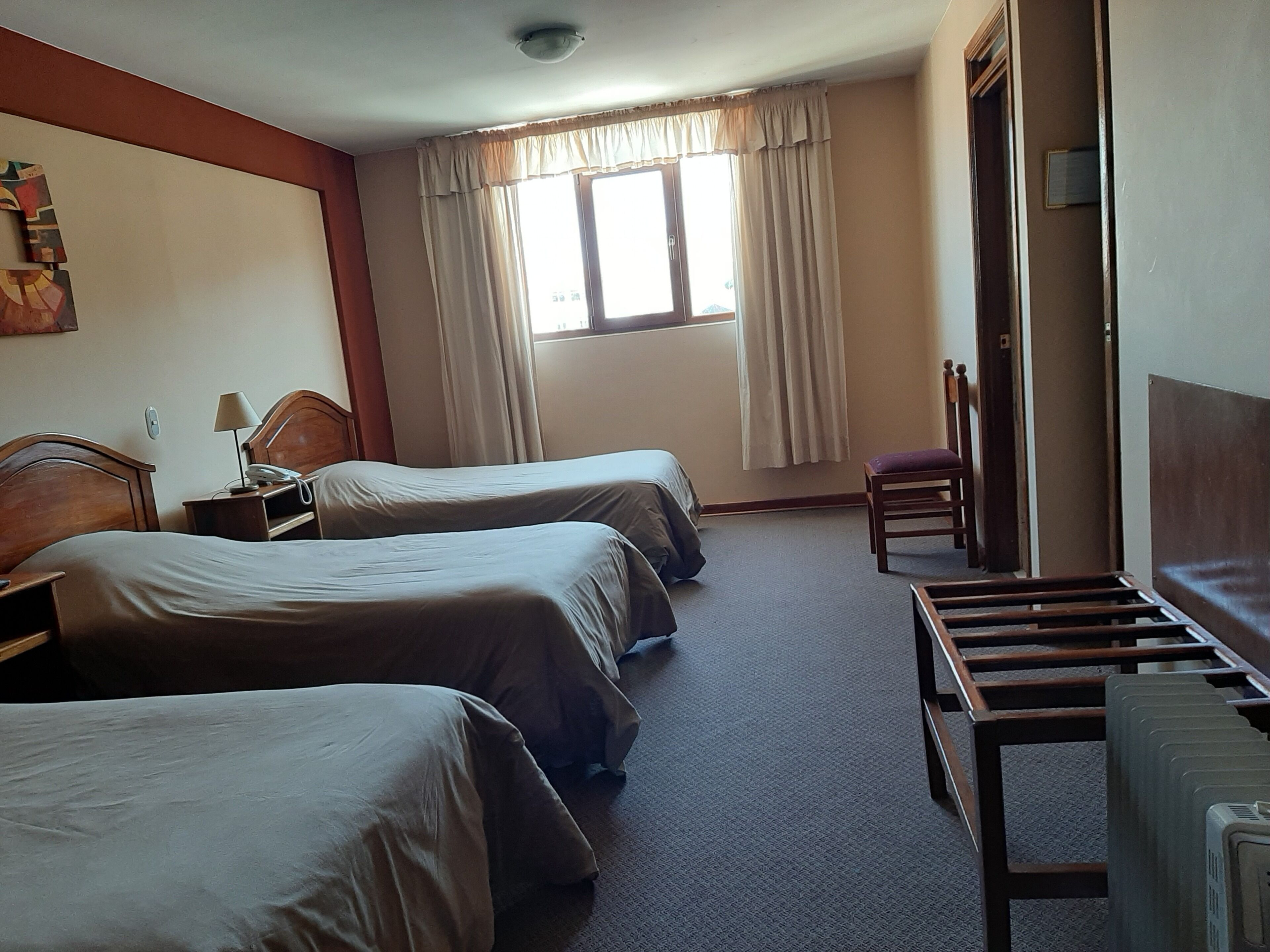 A hotel room with two beds, a desk with a lamp, a chair, and a radiator.