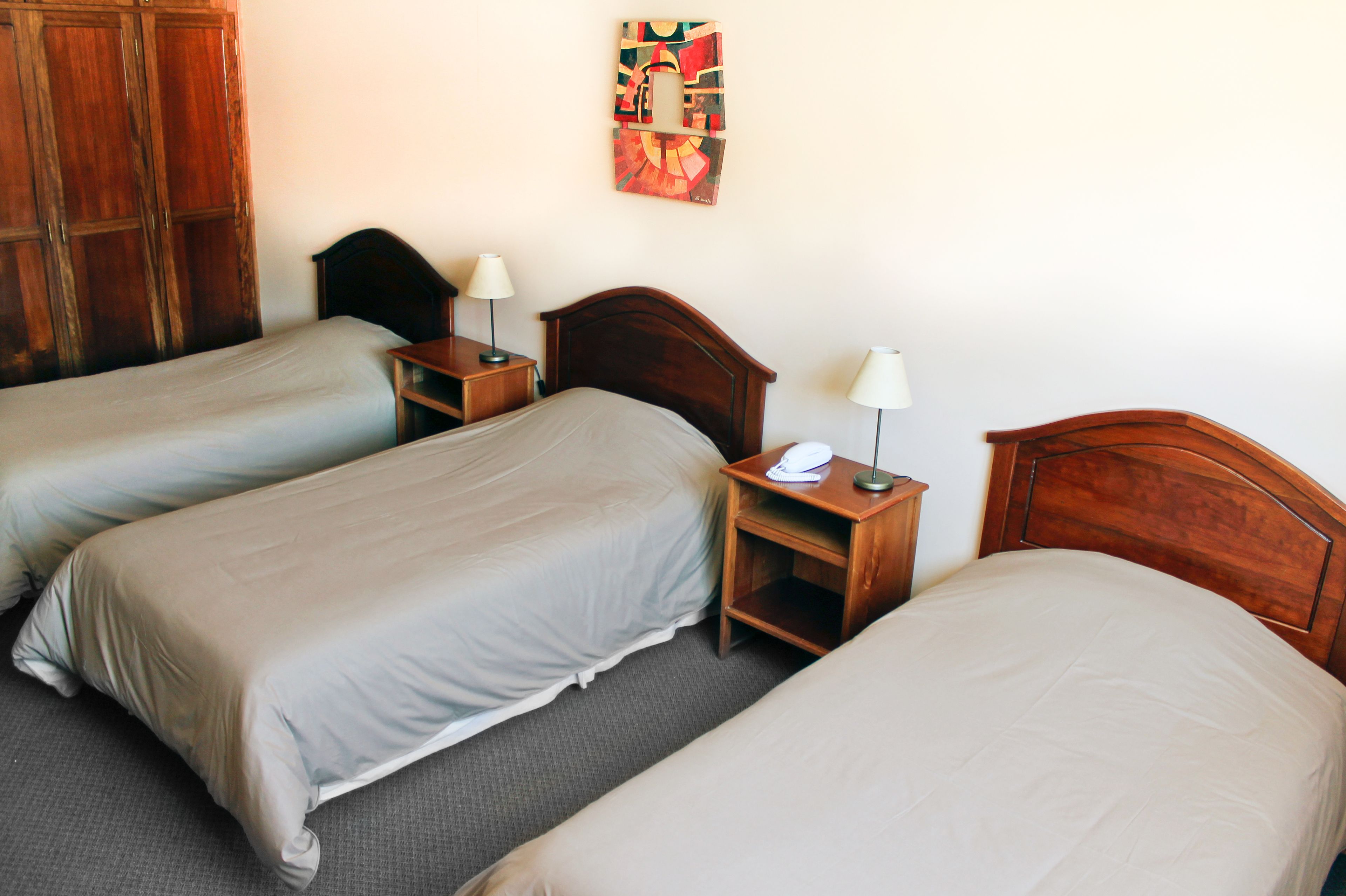 A hotel room with three single beds, wooden headboards, bedside tables with lamps, and a wall-mounted artwork.