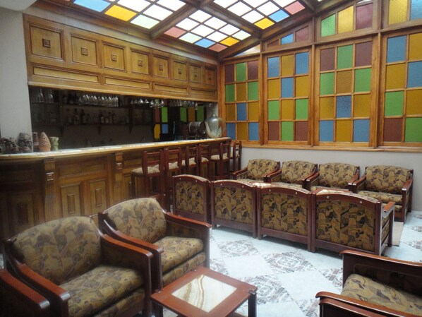 Una zona de bar con armarios de madera y suelo de baldosas. Se observan varias sillas de madera y un sofá con tapicería estampada.