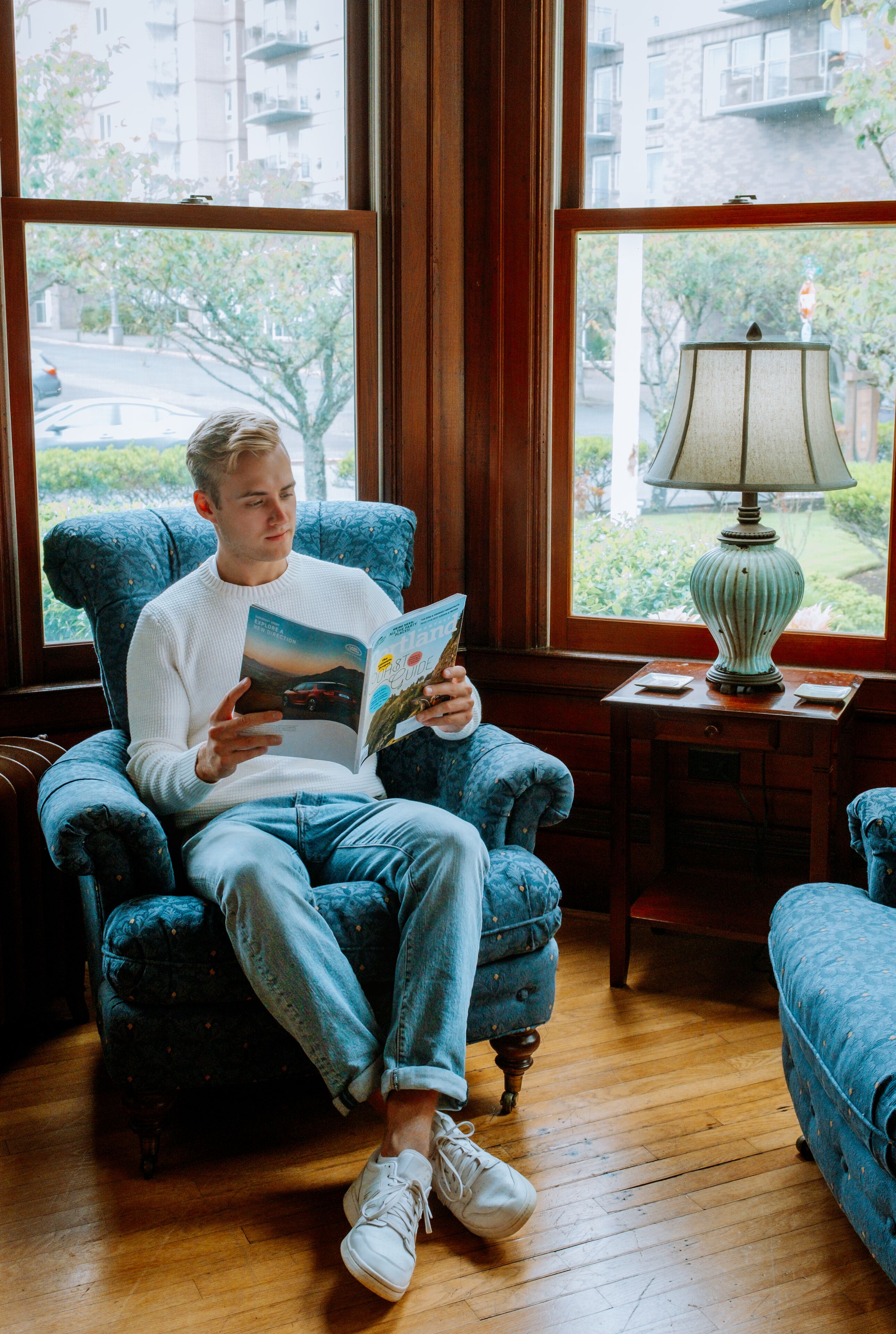 A man sitting in a blue armchair reading a book in a room with wooden floors and large windows.