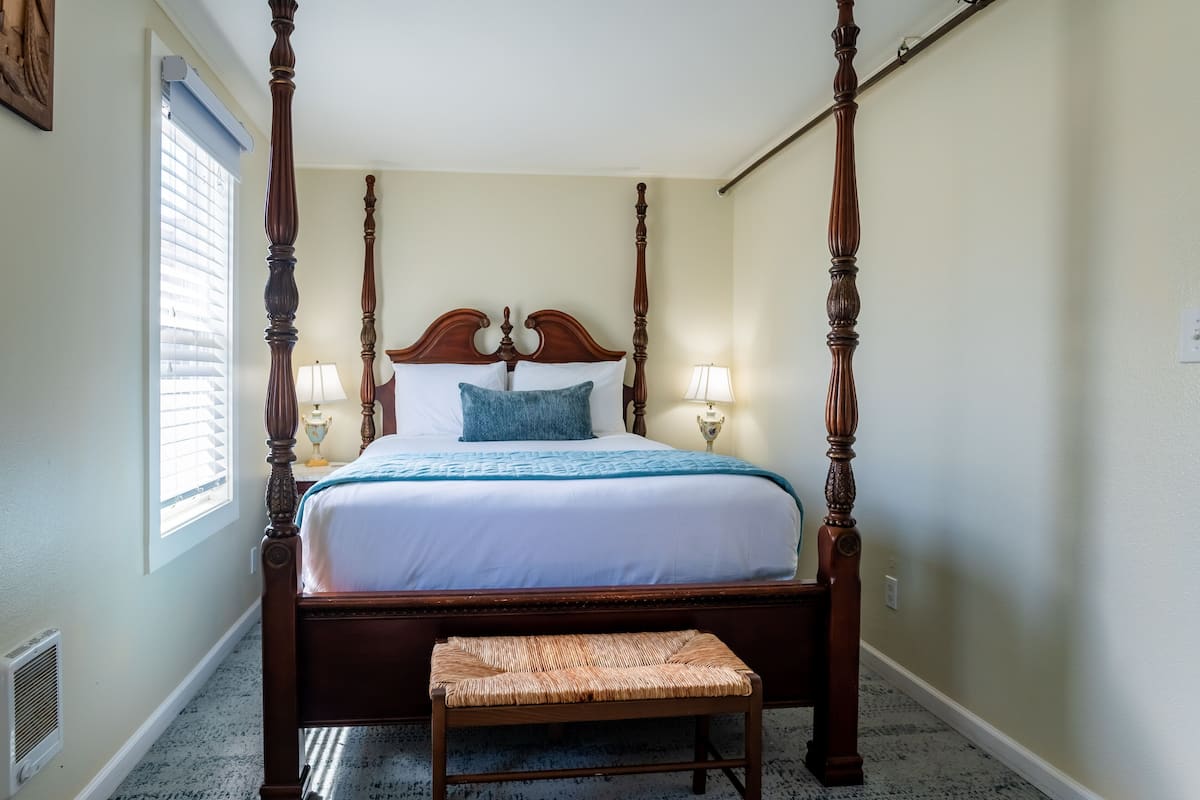 A four-poster bed with a wooden frame, a white bedspread, and two blue pillows. There is a bedside table with a lamp on each side of the bed.