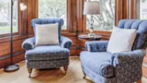 Una habitación con ventanas amplias, un sillón azul, un sofá y una lámpara de pie.