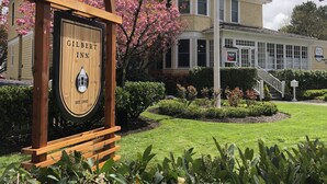 Un letrero de madera del Gilbert Inn con un diseño clásico, un jardín bien cuidado y un edificio tradicional con ventanas grandes y una torre en el fondo.