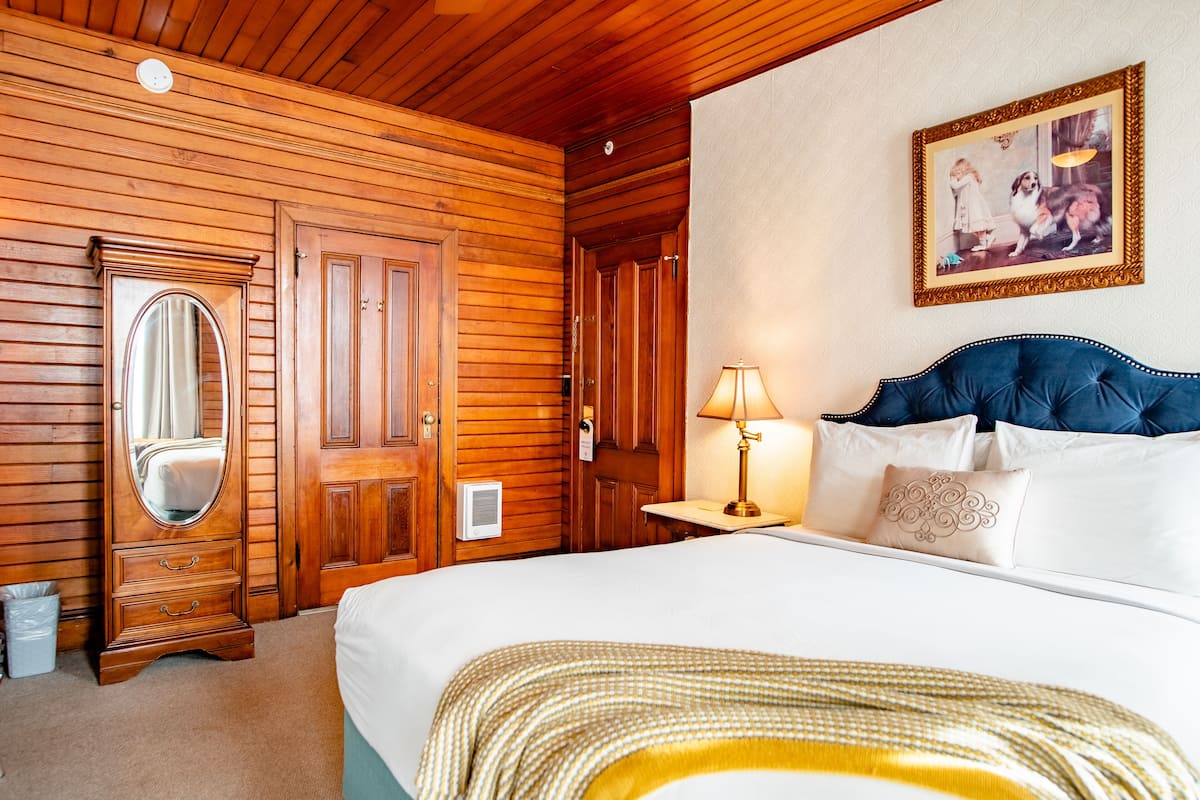 A wooden-paneled room with a bed, a dresser with a mirror, a framed picture on the wall, and a nightstand with a lamp.