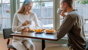 Dos personas sentadas en una mesa de madera con vista a la ventana, un plato de comida y una taza.