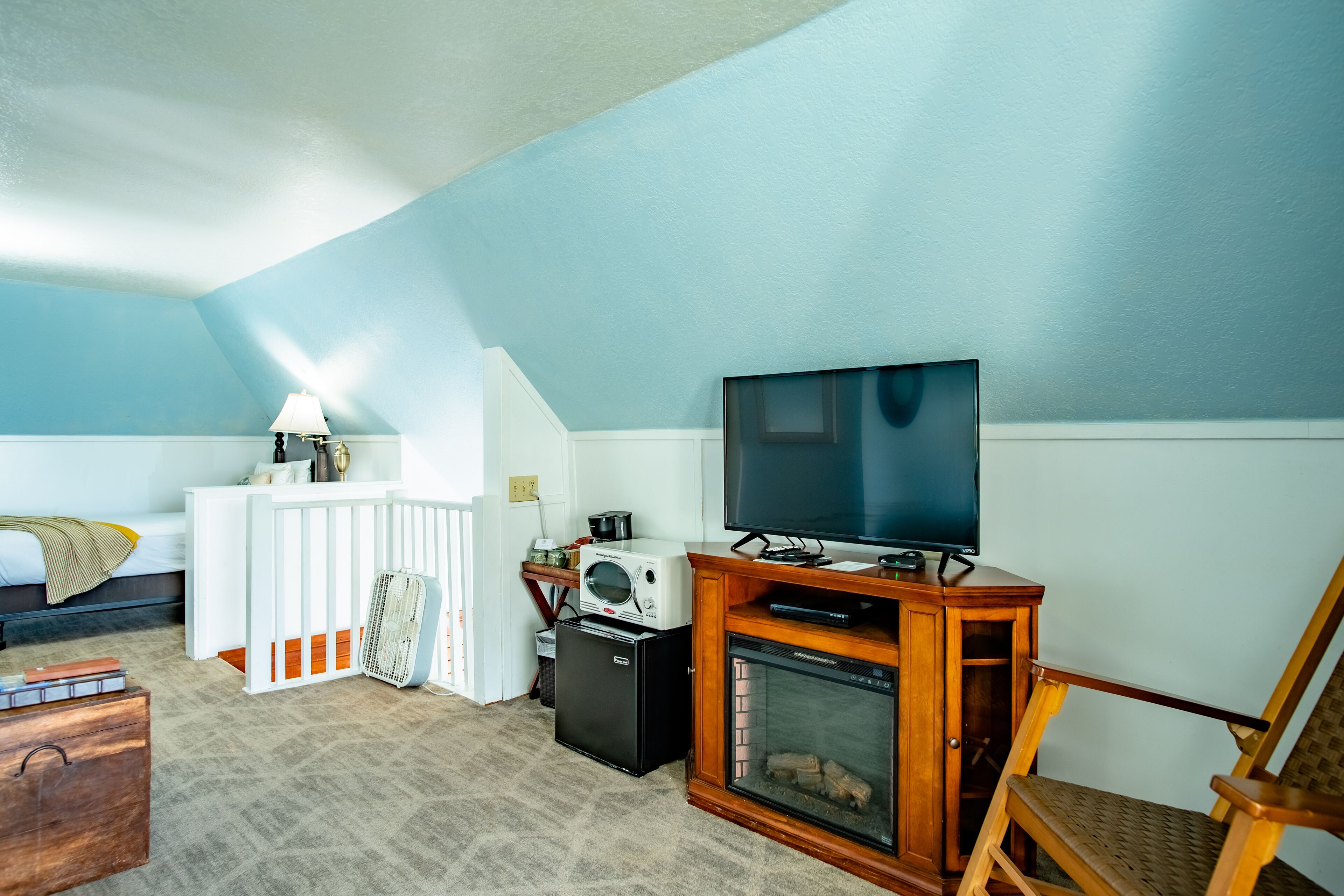 Een knus vertrek met een bed, een wit bureau met lamp, een televisie op een houten standaard, een kleine koelkast en een open haard.