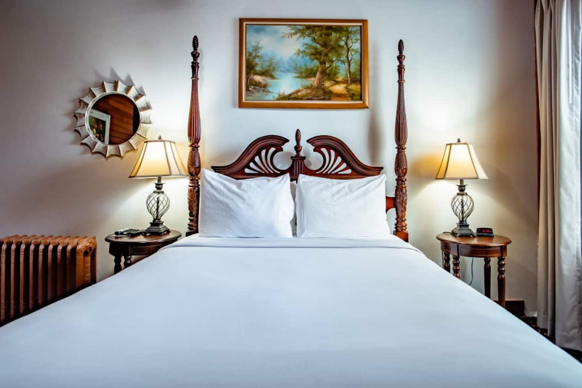 A neatly made bed with a wooden headboard, two bedside tables with lamps, a painting on the wall, and a radiator.