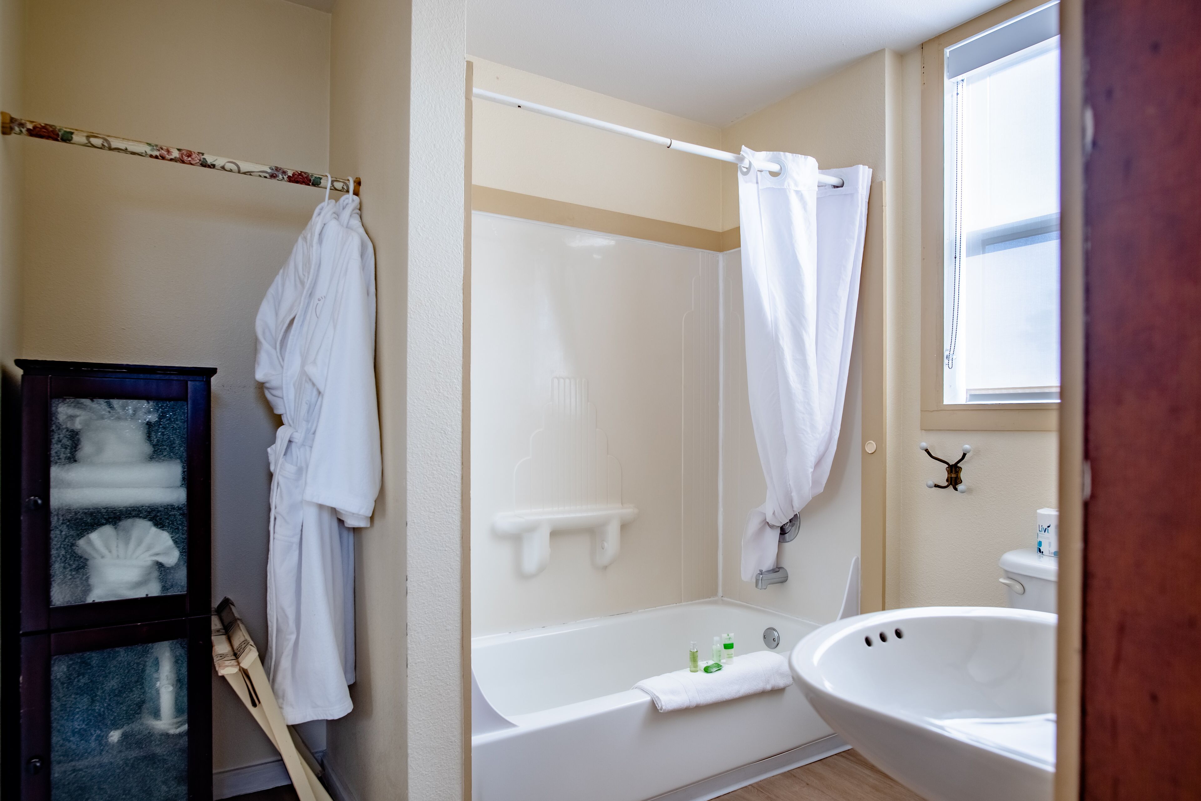Un baño con bañera, lavamanos y ducha. Toallas cuelgan de un perchero y hay un mueble de madera con puertas de cristal.