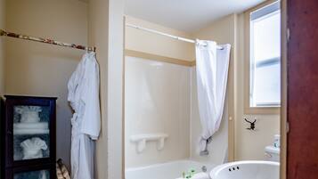 Un baño con bañera, lavamanos y ducha. Toallas cuelgan de un perchero y hay un mueble de madera con puertas de cristal.
