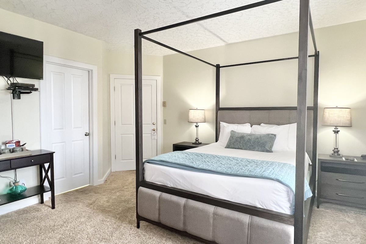 A four-poster bed with a grey upholstered headboard, two bedside tables with lamps, a desk with a lamp, and a white door.