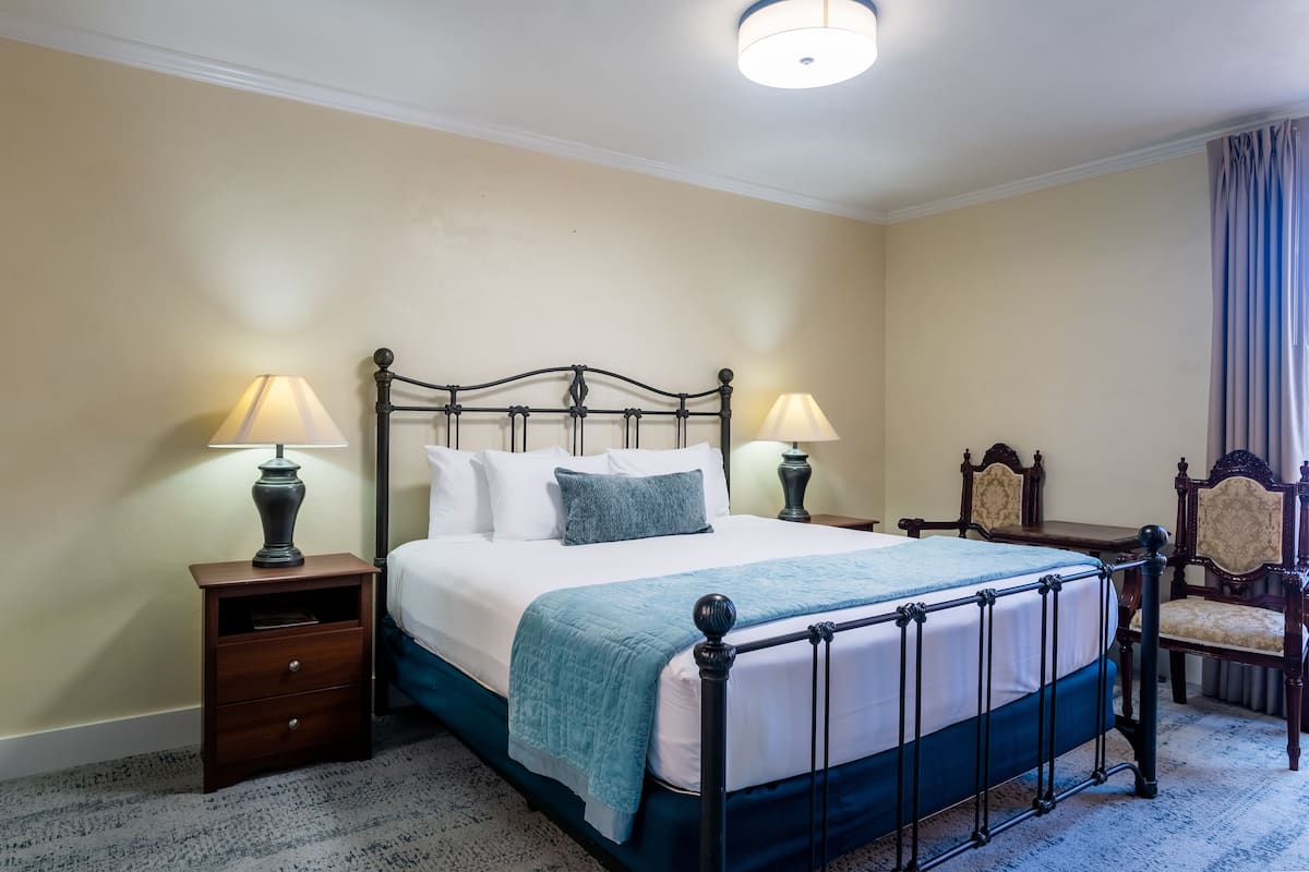 A neatly made bed with a headboard, two bedside tables with lamps, a chair, and a carpeted floor.