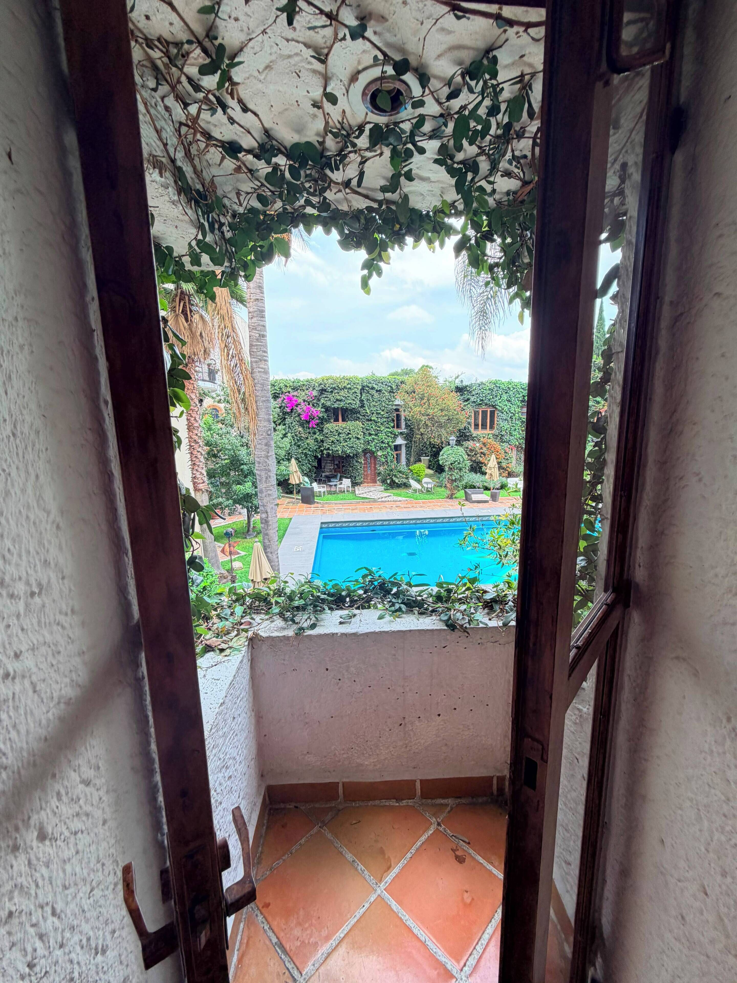 A view through a balcony door of a swimming pool surrounded by greenery and a tiled floor.