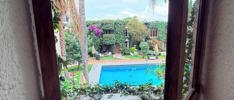 A view through a balcony door of a swimming pool surrounded by greenery and a tiled floor.