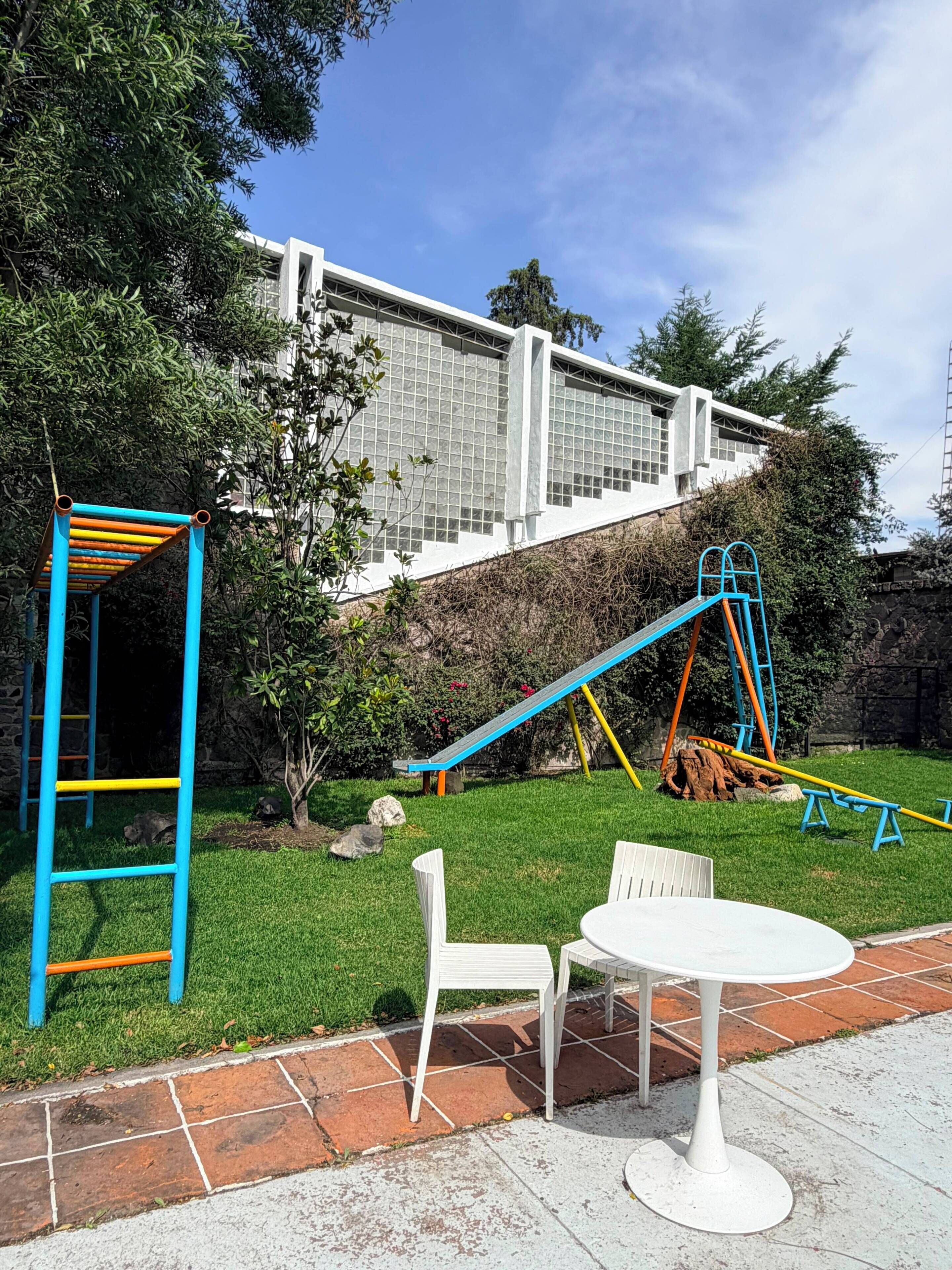 Un parque infantil con un tobogán y estructuras para escalar, una mesa y sillas blancas, y un muro de piedra al fondo.