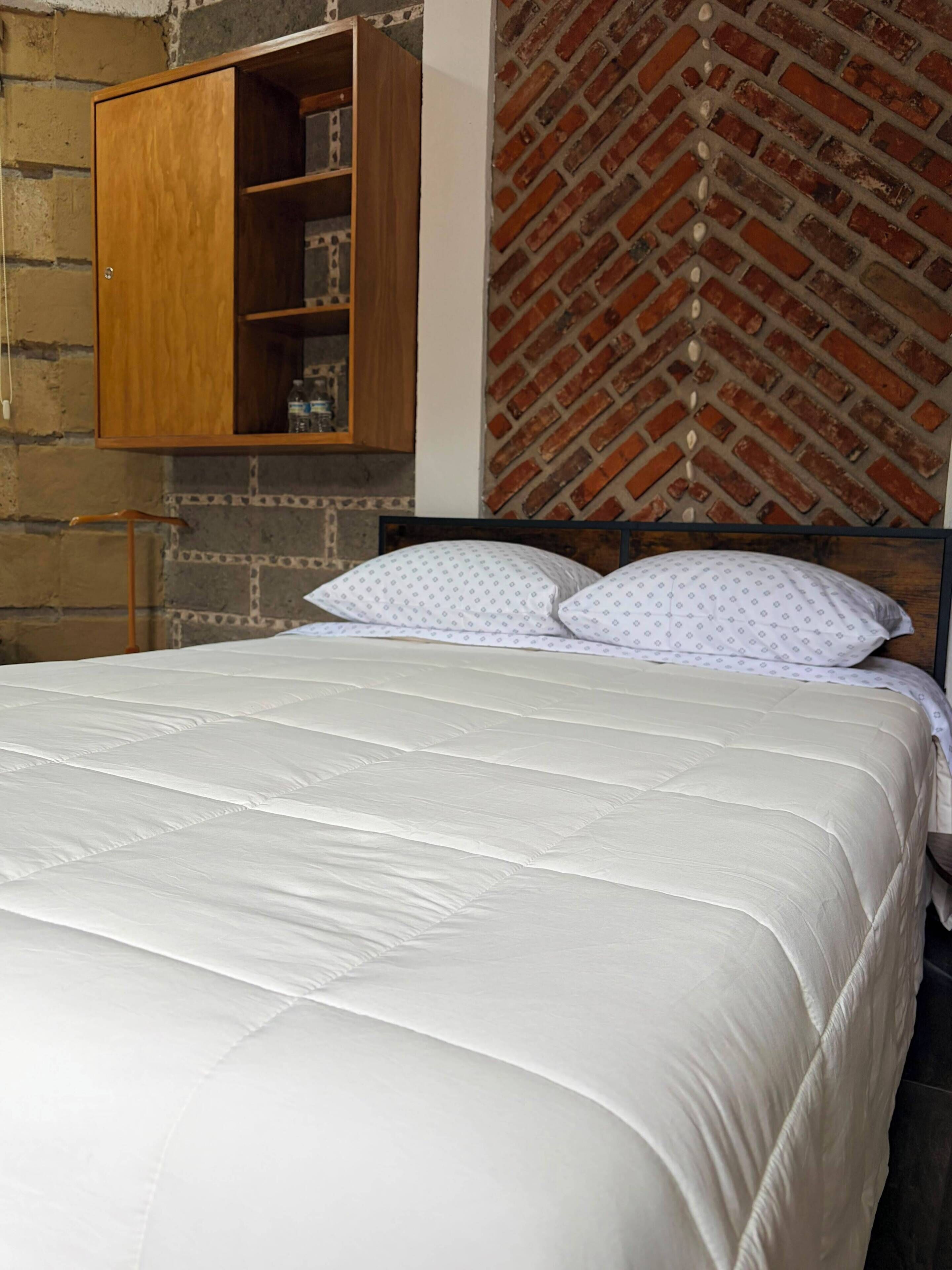 A bed with white bedding and two pillows, a wooden wardrobe, and a brick-patterned wall.