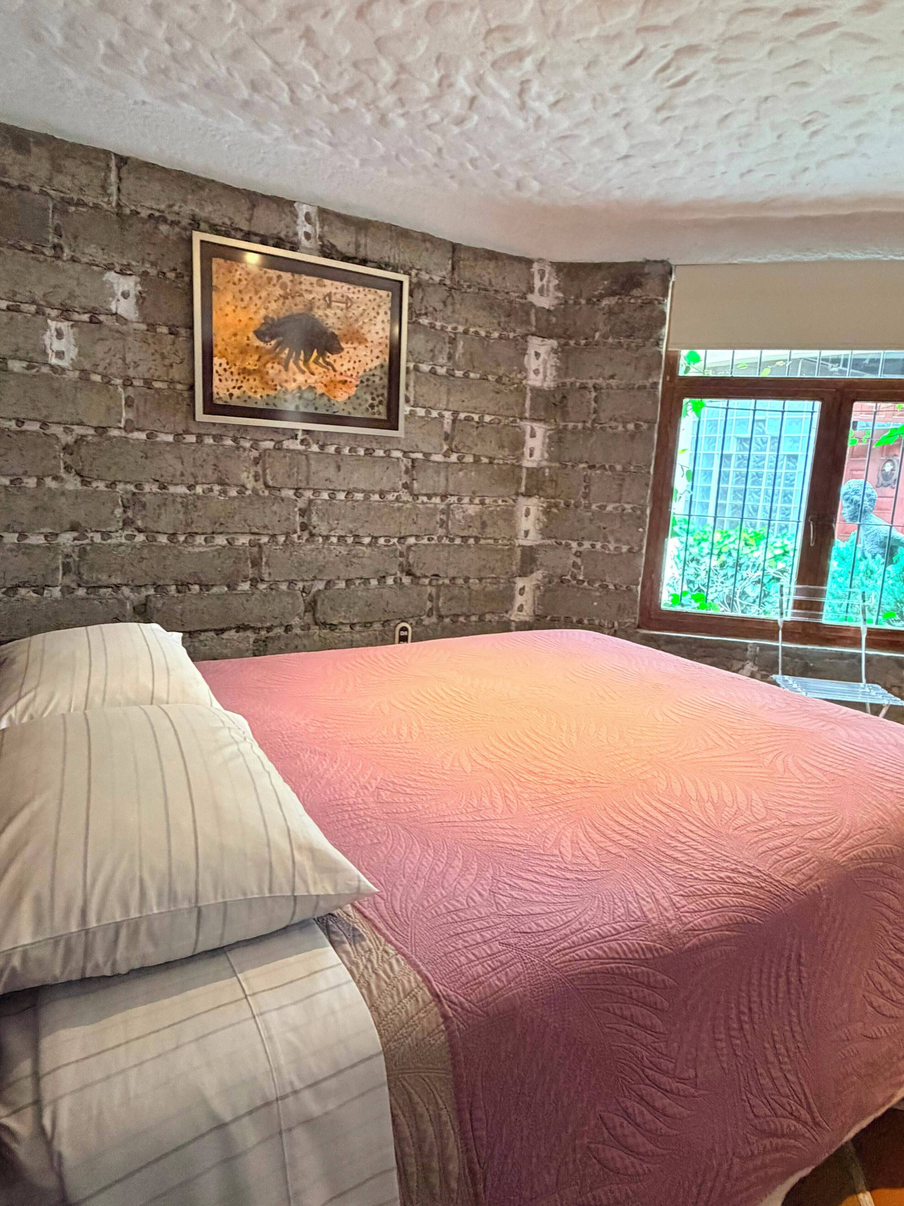 Una habitación con pared de ladrillo, un cuadro enmarcado, una cama con almohadillas y una ventana con vista a follaje.