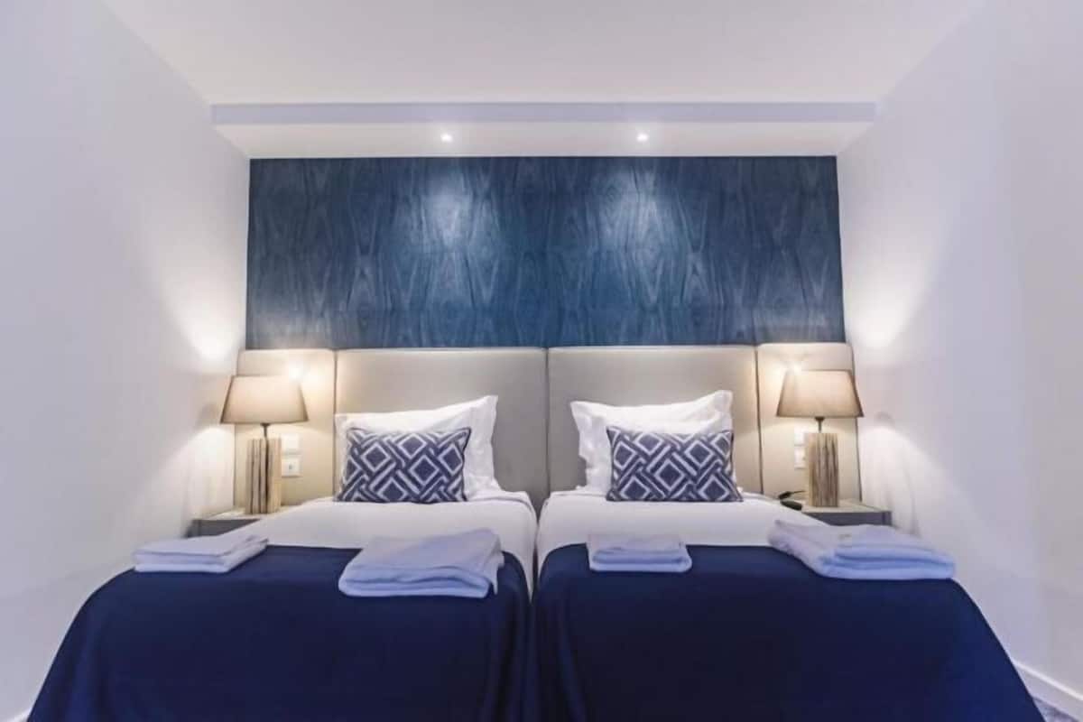 Habitación de hotel con dos camas, cada una con cabecera, lámparas de noche y colcha azul.