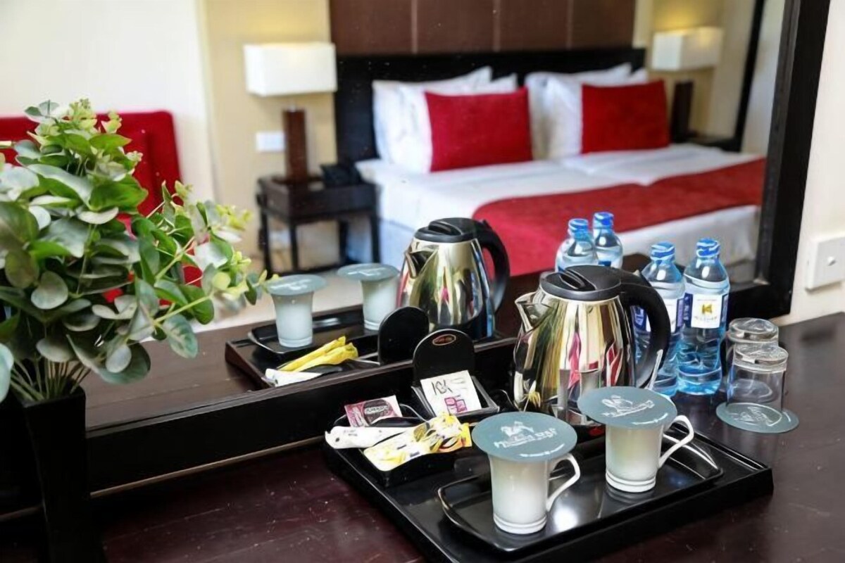 Une chambre d’hôtel avec un lit, une table de chevet, une lampe, un oreiller rouge, une plante et un plateau avec du café et des bouteilles d’eau.