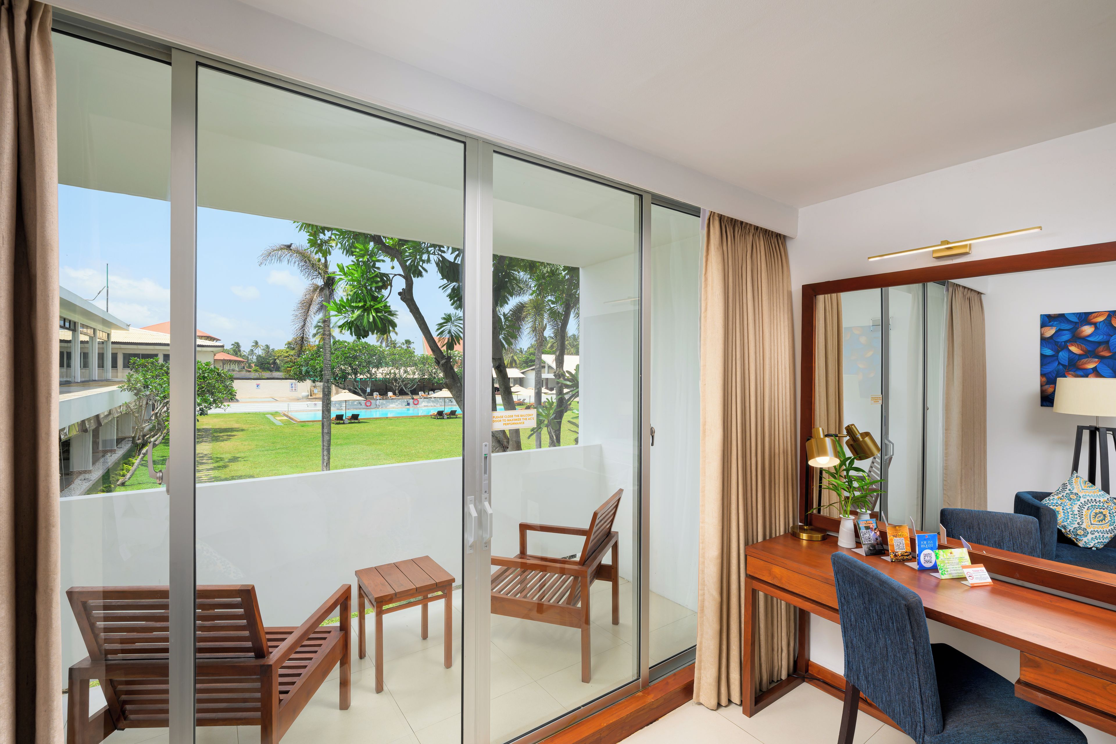 Habitación de hotel con balcón, un escritorio con silla, una lámpara y vistas a una piscina y vegetación.