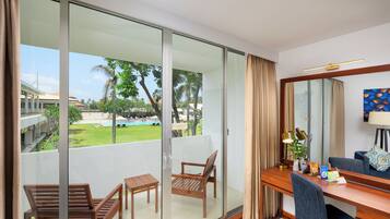 Habitación de hotel con balcón, un escritorio con silla, una lámpara y vistas a una piscina y vegetación.