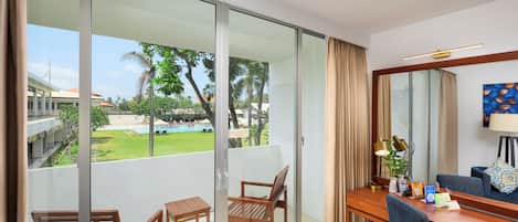 Habitación de hotel con balcón, un escritorio con silla, una lámpara y vistas a una piscina y vegetación.