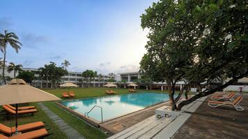 A swimming pool with lounge chairs and umbrellas, surrounded by greenery and buildings.