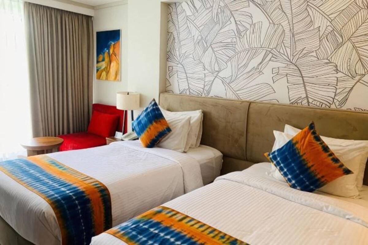 Une chambre d’hôtel avec deux lits, une chaise rouge, une lampe et un papier peint à motifs de feuilles.