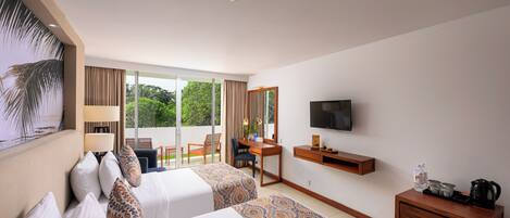 A hotel room with two beds, a desk, a chair, a TV, and a balcony with a view of greenery.