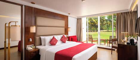 A hotel room with a large bed, a nightstand, a desk with a coffee maker, and a view of a garden through the window.