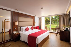 Habitación de hotel con una cama grande, una mesita de noche, un escritorio con cafetera y vistas a un jardín a través de la ventana.