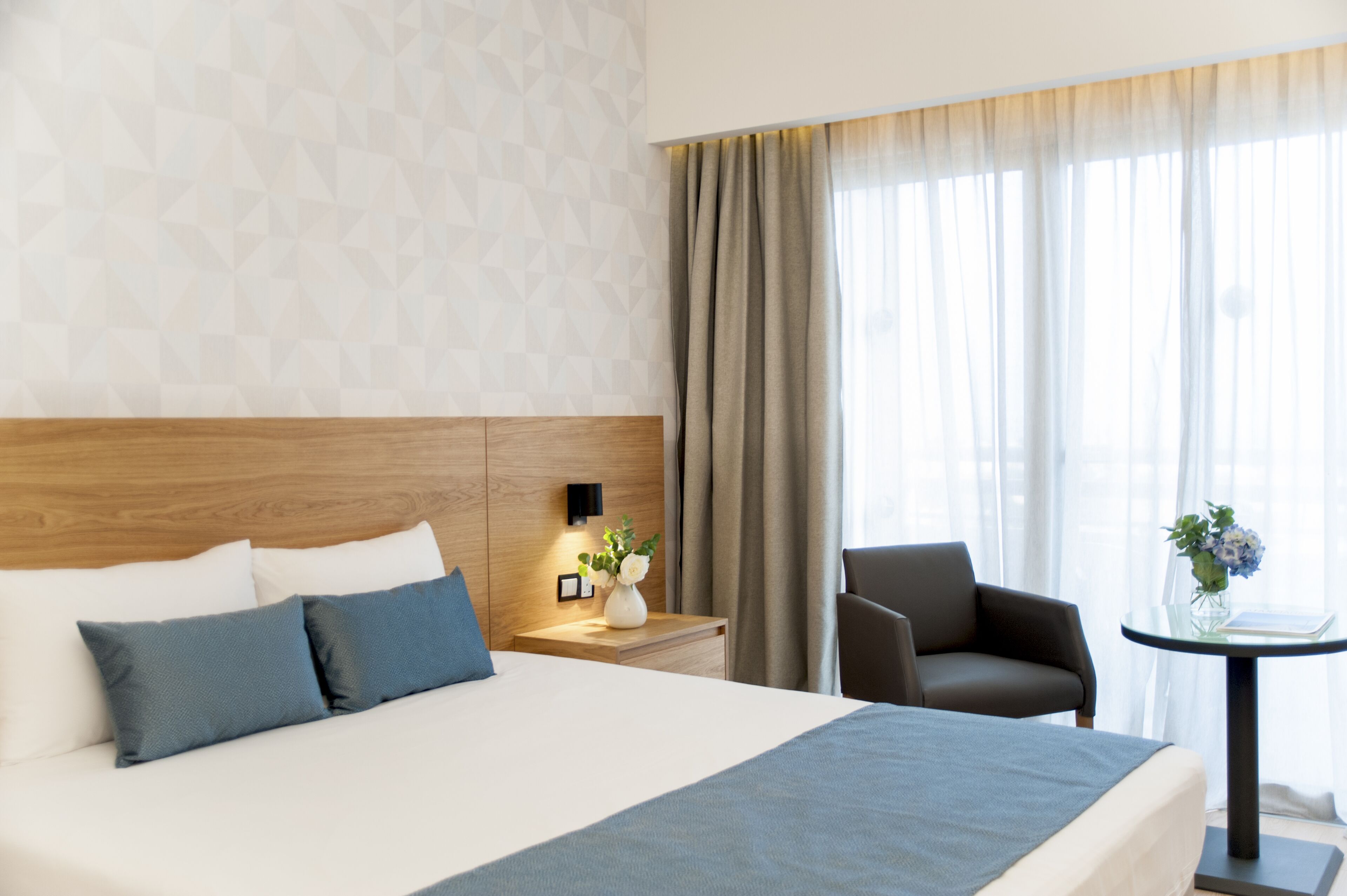 A hotel room with a large bed, a bedside table with a lamp, a chair, and a small table with a vase of flowers.