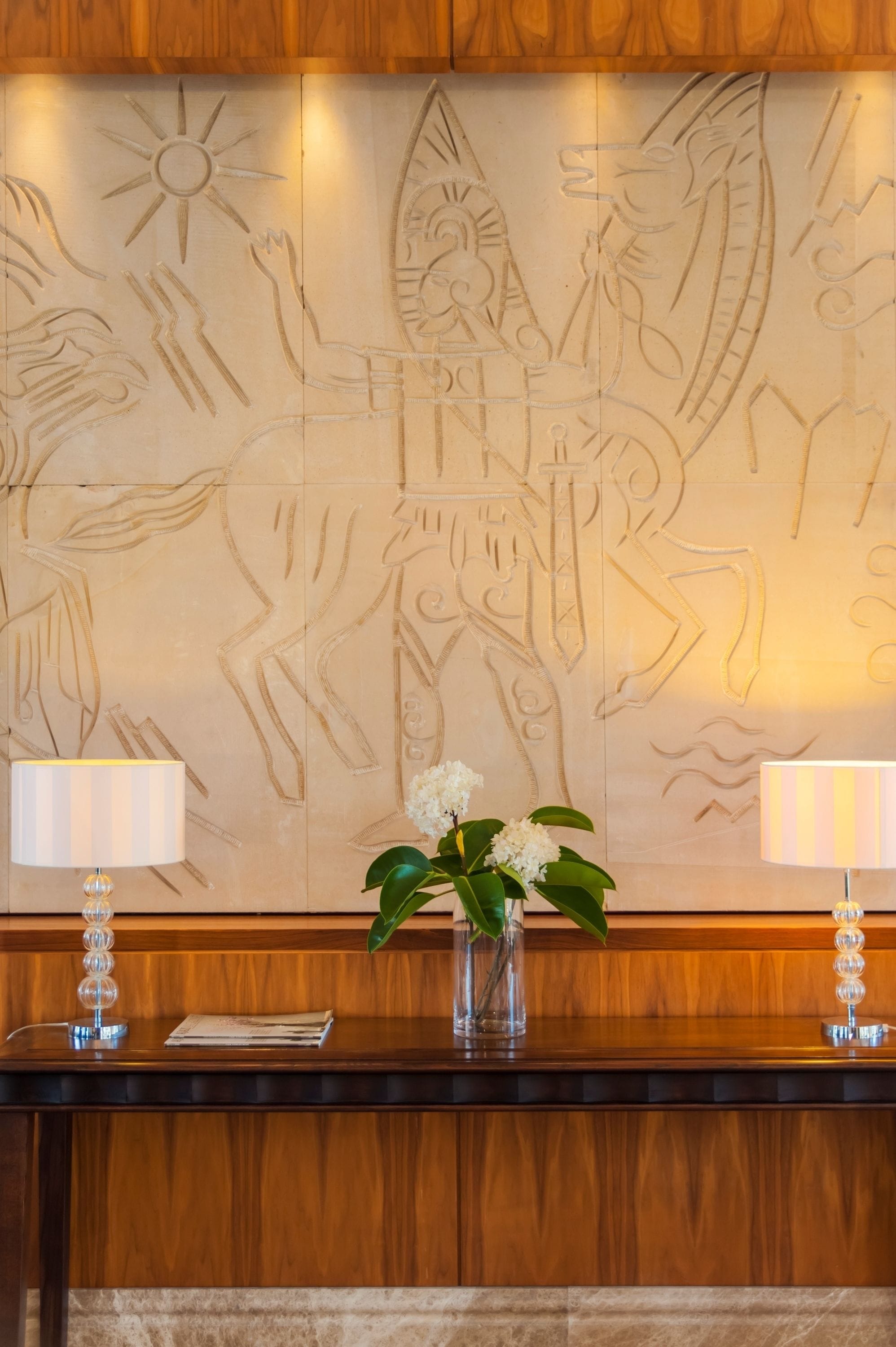A reception desk with a wooden finish, a wall mural with ancient carvings, and two lamps on either side.