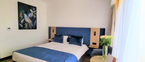 A hotel room with a bed, a nightstand, a wall clock, a painting, a vase with flowers, and a small table with a menu.