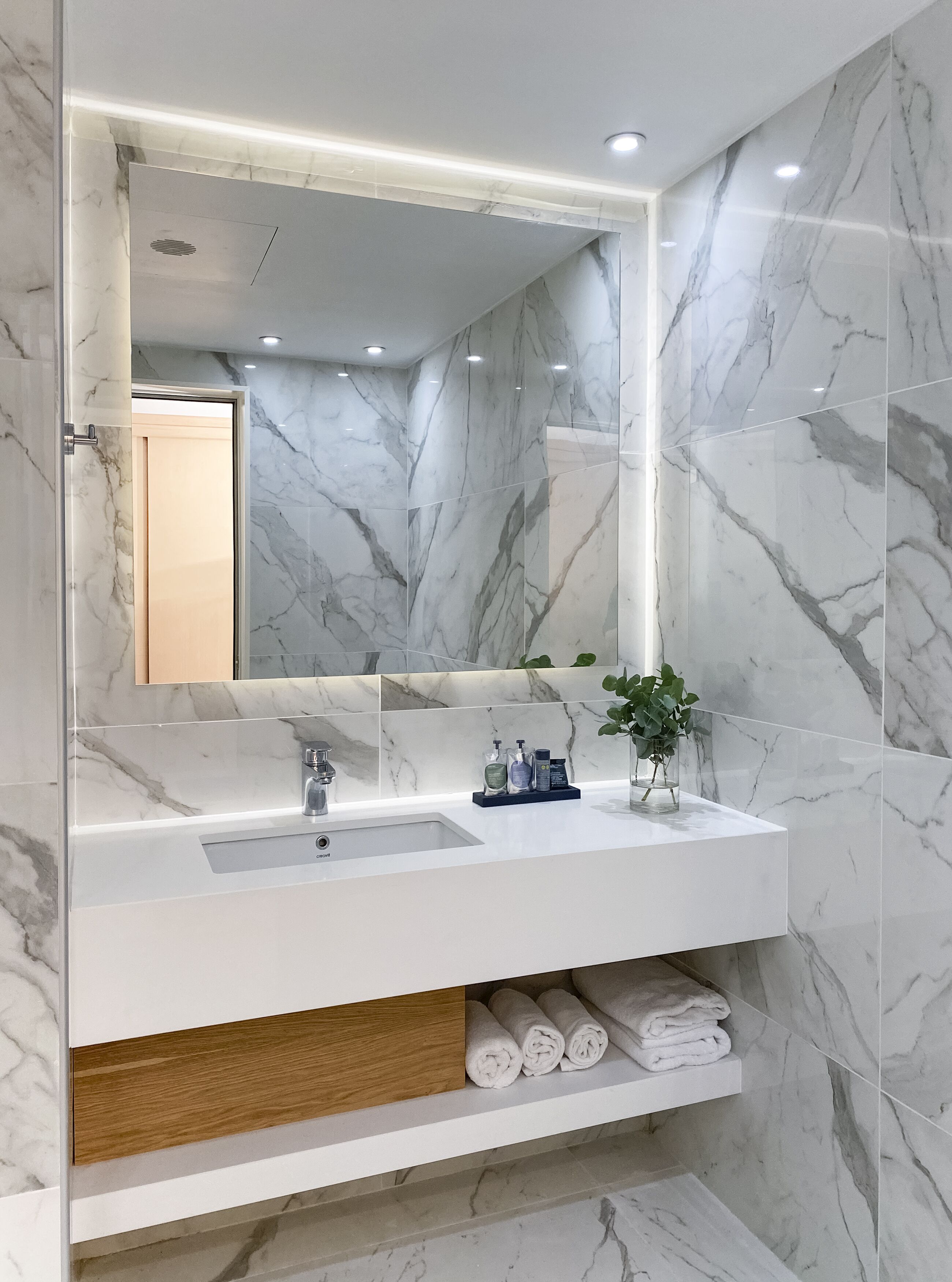 Un baño con tocador de mármol, un espejo grande y repisas con toallas.