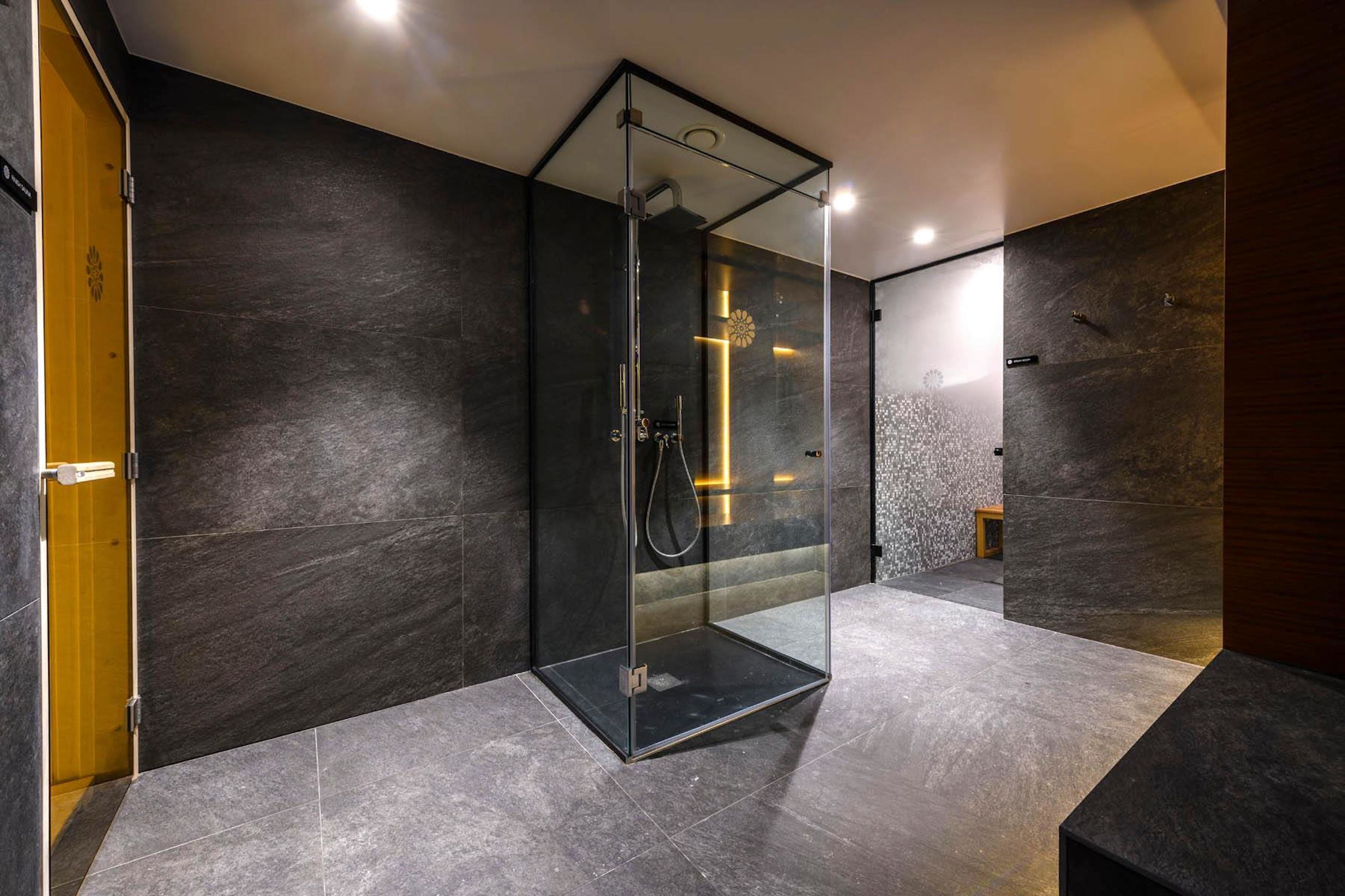 Un baño moderno con cabina de ducha de vidrio, paredes de piedra oscura y un taburete de madera pequeño.