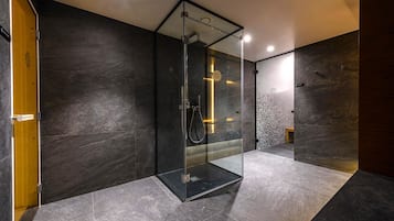 Un baño moderno con cabina de ducha de vidrio, paredes de piedra oscura y un taburete de madera pequeño.