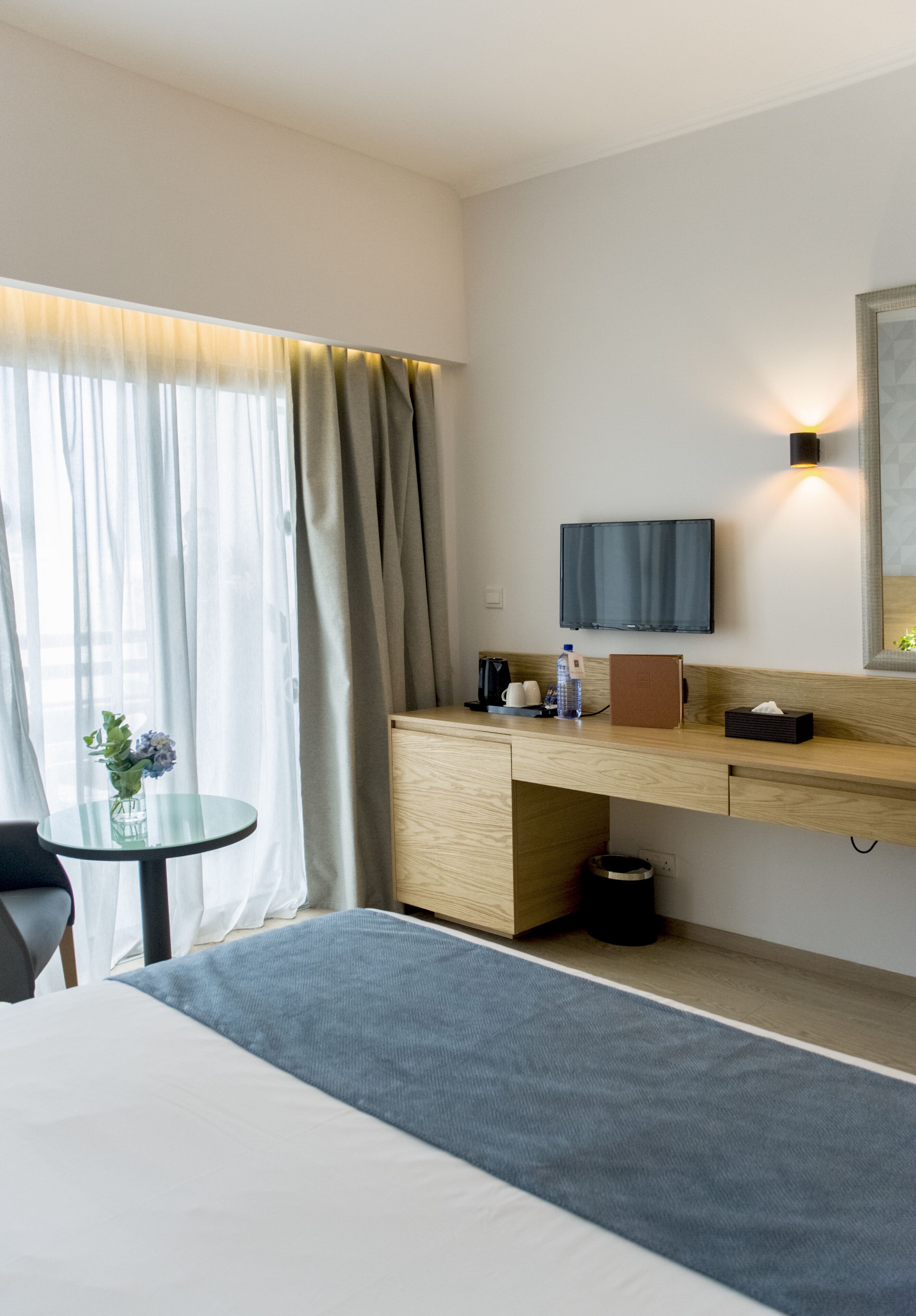 Habitación de hotel con cama, escritorio con computadora, televisor y una mesita con un jarrón de flores.