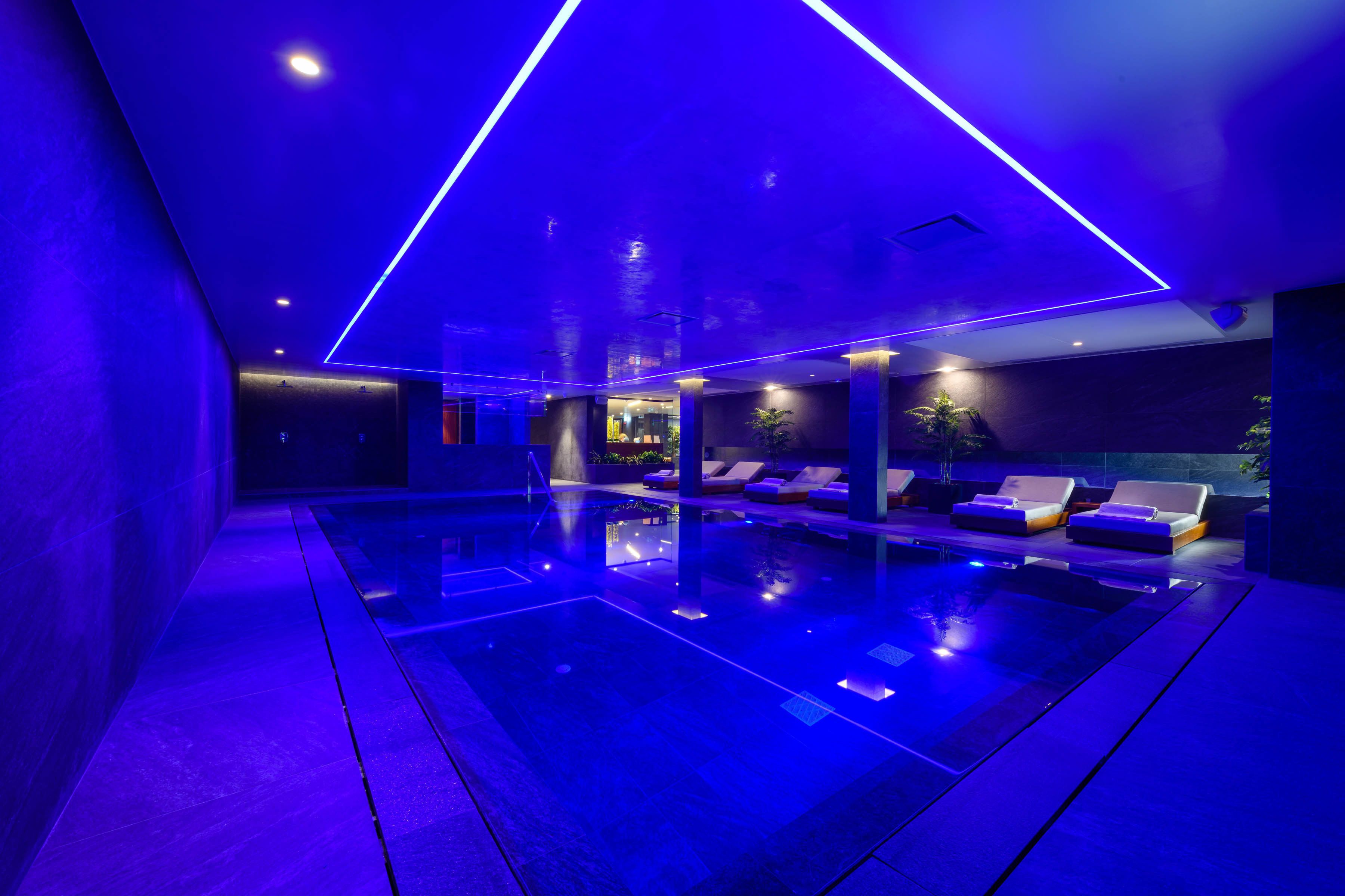 Un área de piscina interior moderna con iluminación azul, sillones de descanso y una barrera de cristal.