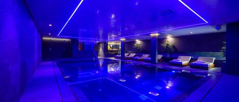 Un área de piscina interior moderna con iluminación azul, sillones de descanso y una barrera de cristal.