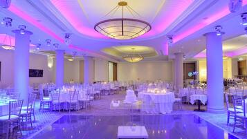A spacious banquet hall with round tables and chairs, a central chandelier, and a polished floor.