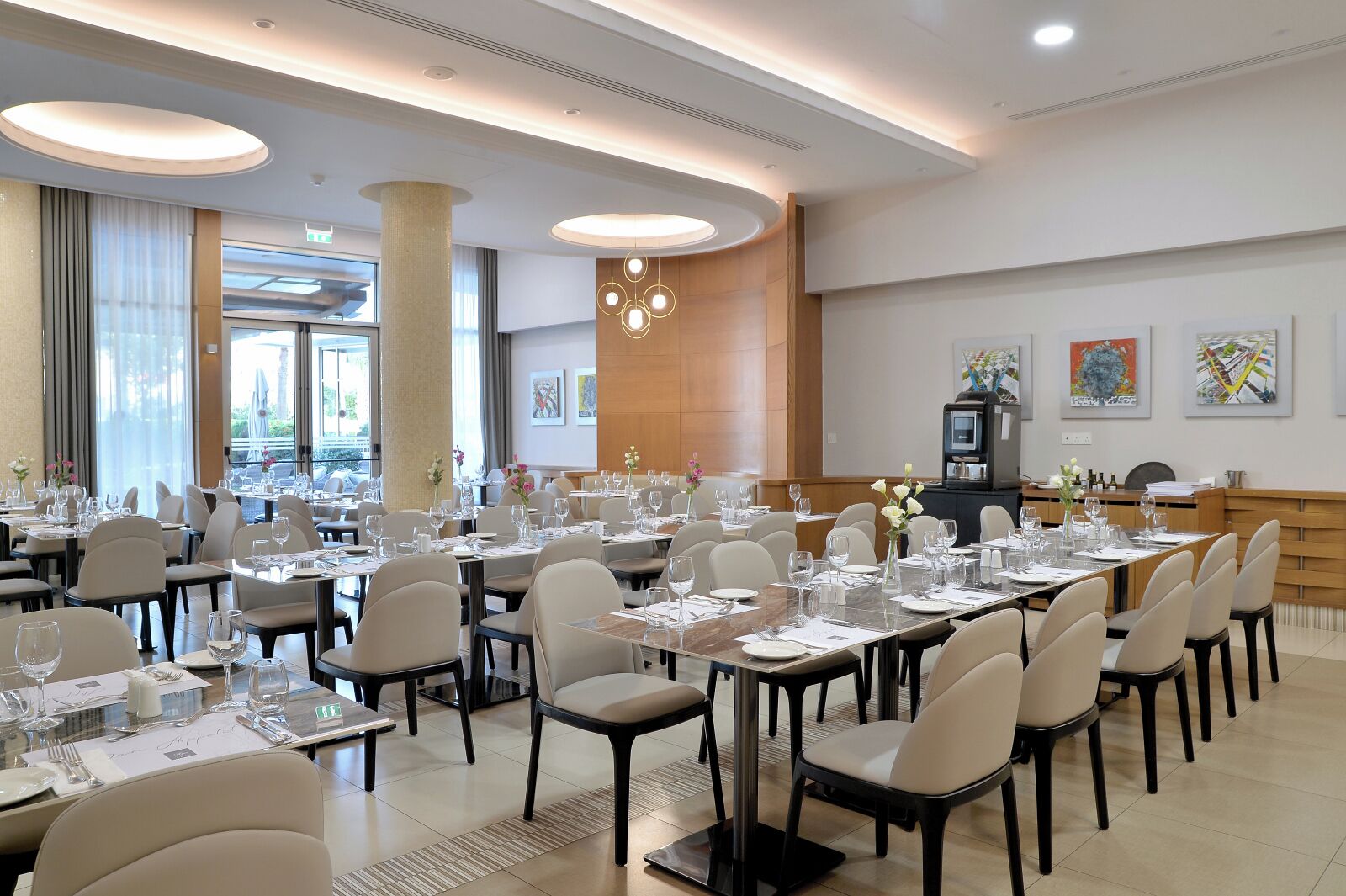 A spacious dining area with multiple tables set for guests, large windows allowing natural light, and artwork on the walls.