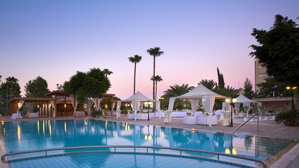 A swimming pool with lounge chairs and tables, surrounded by palm trees and a building in the background. - Ajax Hotel (Limassol)