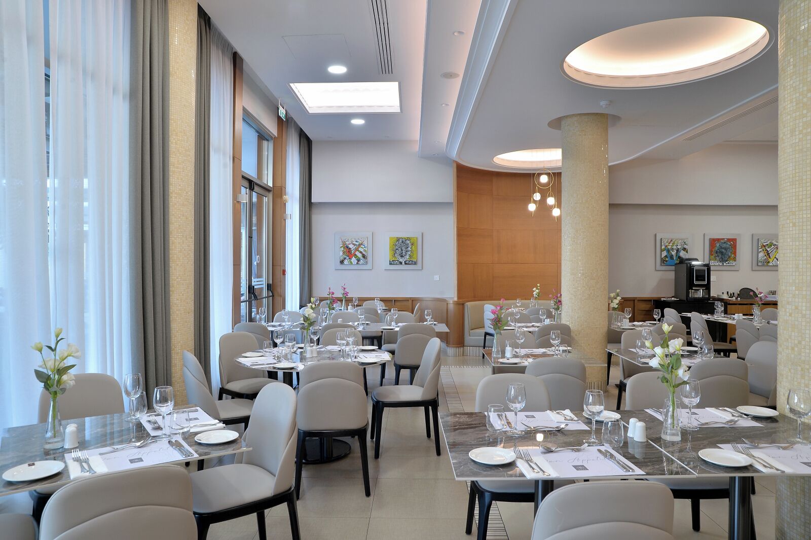 A modern dining area with multiple tables set for guests, large windows, and a kitchen area in the background.