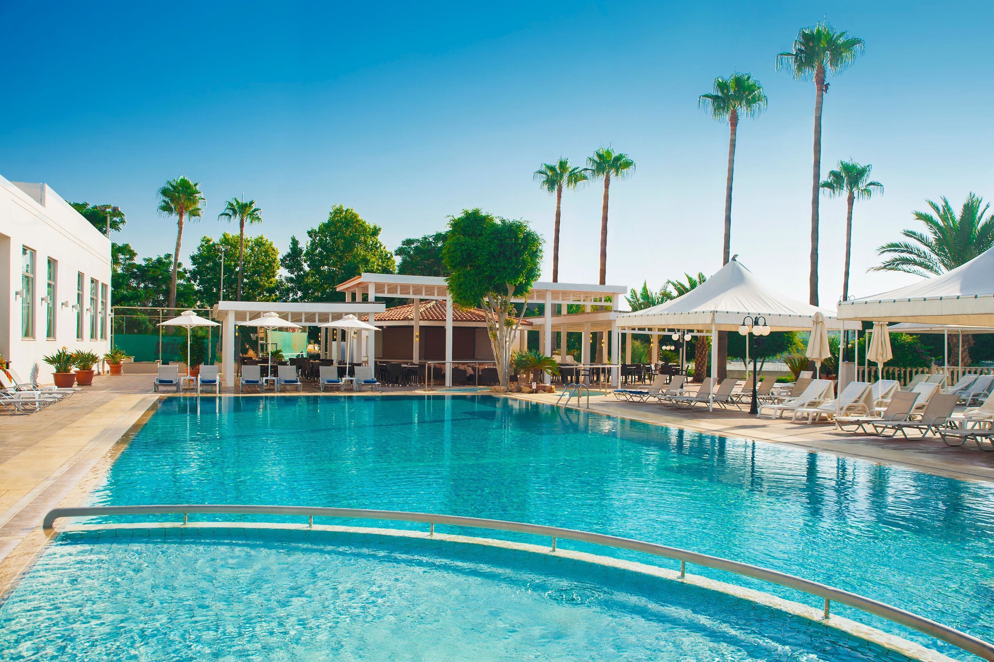 Una piscina con sillones de descanso y sombrillas, rodeada de palmeras y edificios.